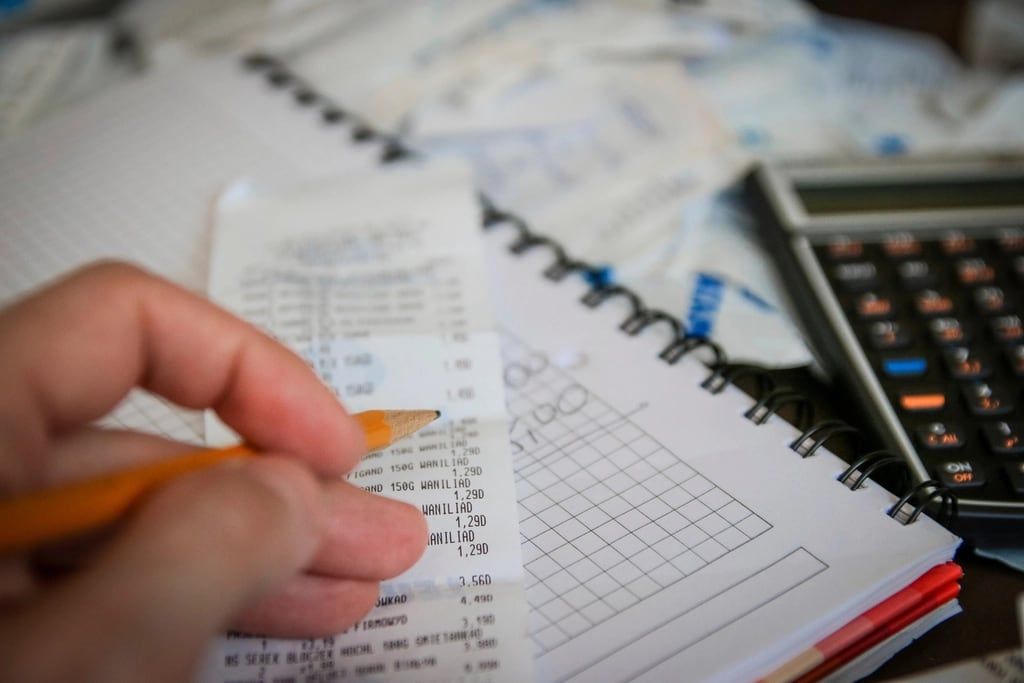 Close-up of a hand with a pencil checking a receipt next to a calculator and a ruled notebook