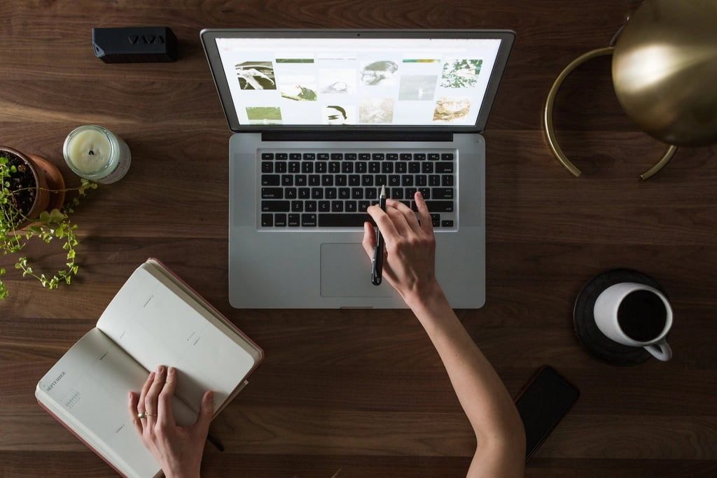 Overhead view of someone working at a laptop with a notebook and coffee on a wooden desk